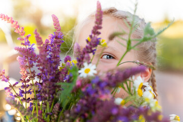 Close-up portrait of little cute caucasian blond kid girl peeking and hiding holding bouquet of salvia sage wild flowers walking at grass meadow outdoors summer day. Young female person wildflowers
