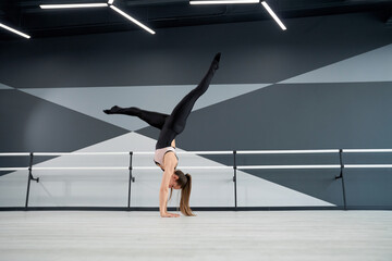 Fototapeta na wymiar Strong fit woman in tight sportswear standing on arms and practicing split in air. Side view of flexible female dancer training before competition in big empty dance hall with ballet handrails.