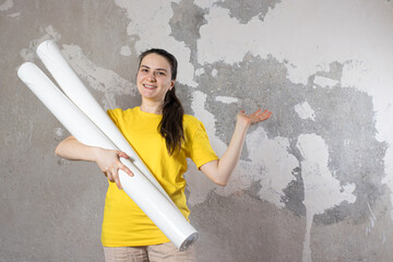 A woman in a bright yellow T-shirt holds 2 rolls of white wallpaper against the old wall. Repair work, wallpapering. With a place for text copy space