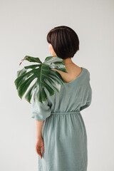 A girl in a menthol-colored dress holds a monstera palm leaf gently from behind. Back photo, no face. Back half-length portrait. Brunette girl. Possibly Chinese, Korean, Japanese, Vietnamese. © Елена Хи