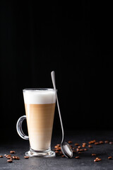 coffee with milk, latte macchiato in a glass with a handle and a long spoon on a black background, vertical