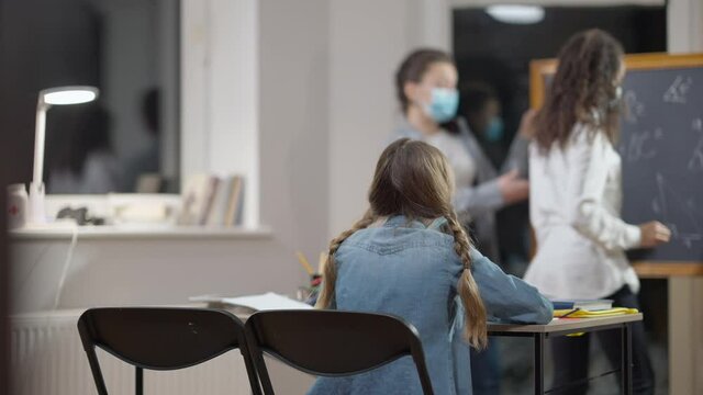 Back view of focused schoolgirl sitting in classroom as blurred African American classmate and Caucasian teacher in Covid face masks talking at chalkboard at background. Coronavirus education concept