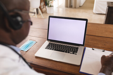 African american senior male doctor writing on clipboard with laptop with copy space on table