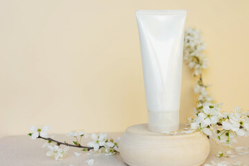 white cosmetic tube for cream on a wooden podium on a beige background with sakura flowers. Gentle skin care concept