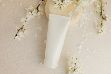 white cosmetic tube for cream on a wooden podium on a beige background with sakura flowers. Gentle skin care concept