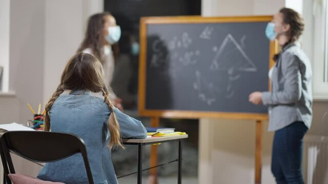 Smart diligent Caucasian schoolgirl sitting at desk raising hand as blurred African American classmate in Covid face mask answering teacher question at chalkboard. Covid-19 studying in school