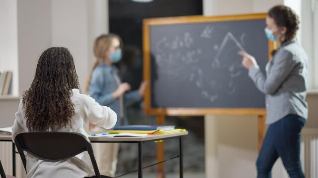 Active African American schoolgirl in Covid-19 face mask raising hand answering teacher question in classroom with blurred Caucasian classmate standing at chalkboard at background