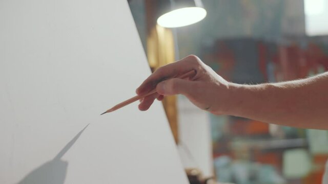 Close up shot of hand of male artist drawing with pencil on blank canvas while working in creative studio with burning incense stick