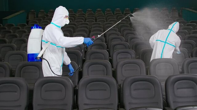 Two men in white protective suit cleaning modern cinema by spraying disinfectant from special equipment. Male cleaners preparing seats for visitors during pandemic time.