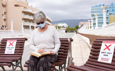 Fototapeta na wymiar Social distancing concept. Senior woman sitting alone on wooden bench reading a book