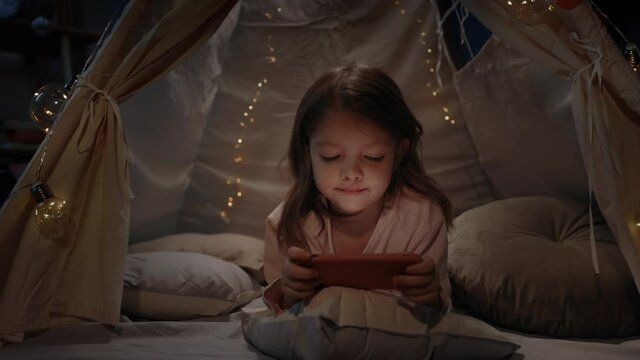 Adorable girl in cute decorative tent playing online games at home in evening. Little kid lying on floor while using smartphone. Concept of leisure and careless childhood.