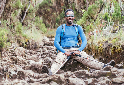 Portrait of a cheerfully smiling African-American Ethnicity young man in sunglasses sitting with backpack and trekking poles and resting on mountain trekking path. Active people and traveling concept