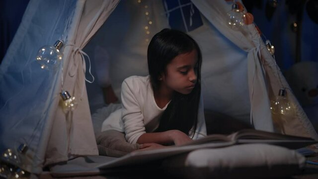Cutre teen girl in decorative tent turning page while reading book in evening. Beautiful teenager lying on floor while spending free time. Concept of leisure and careless childhood.
