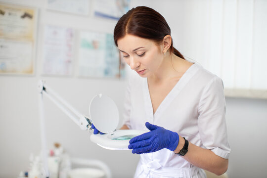 Professional skin problems. Close up portrait of pretty confident female cosmetologist in white uniform and blue protective gloves, looking through magnifying lamp, working at modern clinic