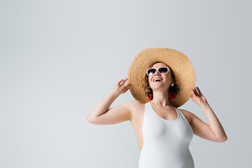 overweight and happy woman in sunglasses and swimsuit adjusting straw hat isolated on white