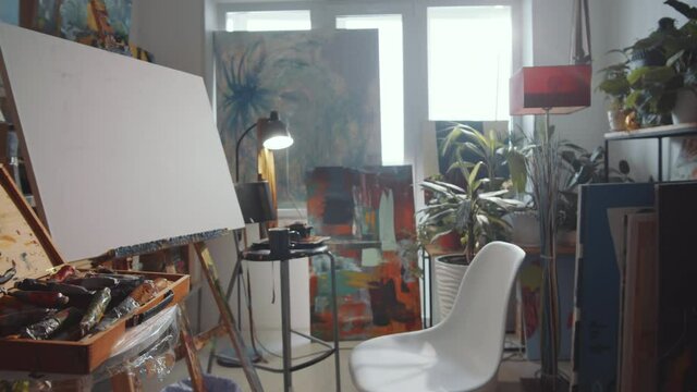 Tracking shot of interior of creative studio of artist with no people inside. There is canvas on easel, chair, case with tools, burning incense stick and paintings