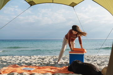 View from inside the tent of young woman in a camping on the beach opening the fridge. Windy and...