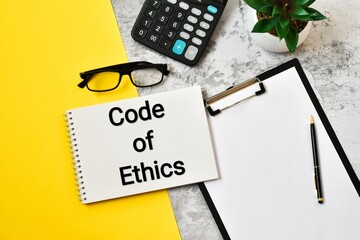 CODE OF ETHICS is written in a white notepad near a clipboard, calculator, green plant, glasses and...