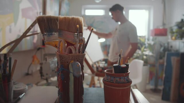 Close up tracking shot of handmade vases with painting brushes, pencils and drawing tools in creative art studio; male artist working on picture in defocused background