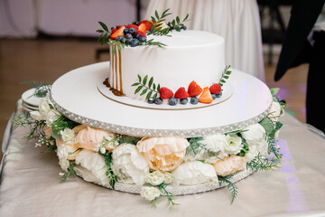 Obraz na płótnie Canvas A beautiful wedding cake, decorated with fruits, raspberries, strawberries and blueberries. The bottom of the cake is decorated with rose flowers. Confectionery on the table. Holiday concept.