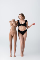 full length of overweight and smiling woman in swimsuit near plastic mannequin on white