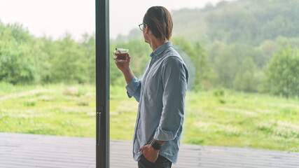 Side view of young businessman in glasses holding cup of coffee and standing near the window