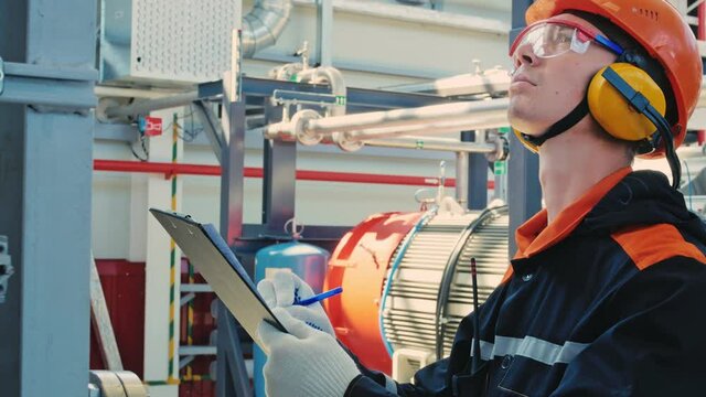 Engineer technician in hard hat, goggles and earmuffs, recording data on the operation of the engine and compressor station. Work in industrial. Manufacturing industry.