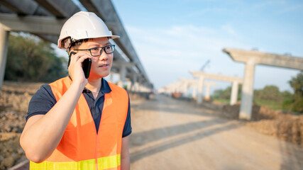 Asian worker man or male civil engineer with protective safety helmet and reflective vest using smartphone for project management at construction site.