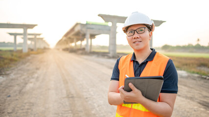 Smart Asian worker man or male civil engineer with protective safety helmet and reflective vest using digital tablet for project planning and checking architectural drawing at construction site.