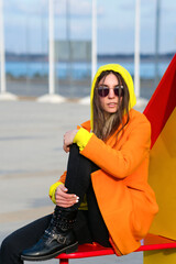 Millennial Hipster Girl in lilac sunglasses, orange coat, yellow hoodie posing at urban background. Trendy colorful casual outfit. Street Style. Fashion lookbook concept