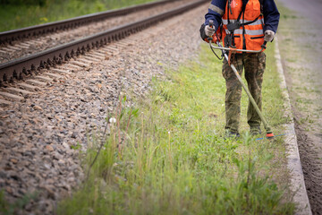 A front view of a worker in protective clothing walks along the lawn next to the train tracks and mows the grass with a gasoline lawn mower.