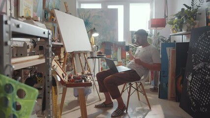 Zoom in shot of young male artist sitting in creative studio, waving, talking and showing painting while video calling on laptop