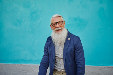 Older caucasian hipster man with prescription glasses and beard smiles happy outdoors in the city - Focus on face.