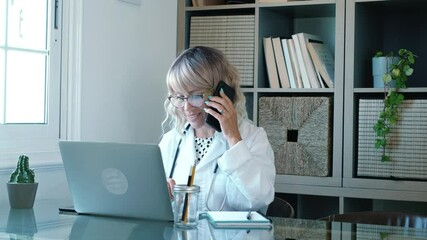 Female adult woman general practitioner wears white coat speaking at the phone and on laptop computer using online video call consultation app. Remote medical help for distance patient
