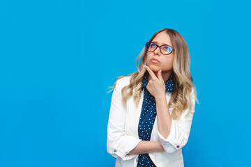 A young caucasian thoughtful pretty blonde woman in a shirt, white jacket and glasses thinks looking up at empty copy space for ads promo offer quote text slogan on a bright color blue background