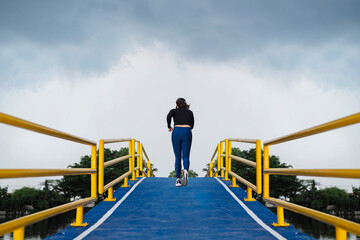 A young woman runs up a hill on a pedestrian bridge over a canal, a blue asphalt road, a yellow...