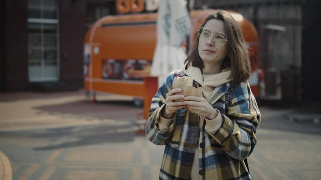 Close Up Of A Millenial Woman In Round Glasses And Yellow Shirt Eating Sandwich On The Street. Foodtruck At The Background. Brunette Woman Having An Hamburger In The Streets