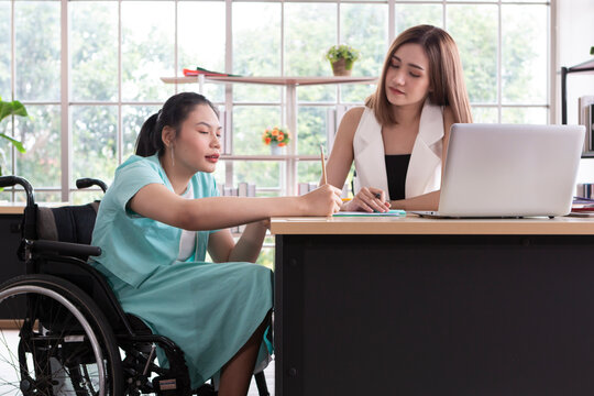 Young disabled woman sitting on wheelchair using computer to discuss project with her female colleagues. Business people on wheelchair in the office. Business Woman talking with disabled colleague.