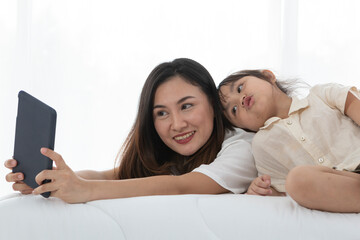 Fototapeta premium Young little girl playing with her mother on bed, she is lying on her mother back looking at tablet screen. Young mother and daughter talking on bed while playing with computer tablet.