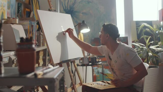 Zoom in shot of talented young man sitting in art studio, holding wooden palette and painting picture on canvas with brush