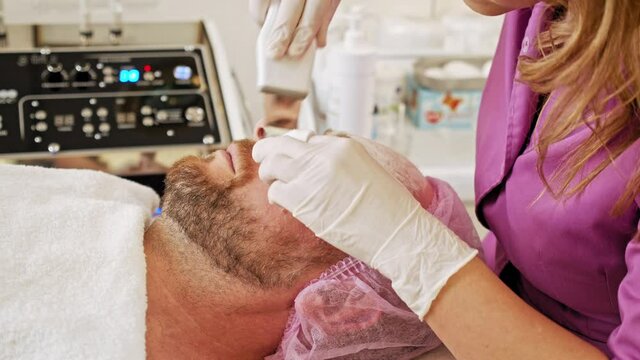 Beautician using electrical impulses for facial procedures.  Man in a spa salon on cosmetic procedures for facial care. Adult man receiving facial microcurrent treatment at spa salon.