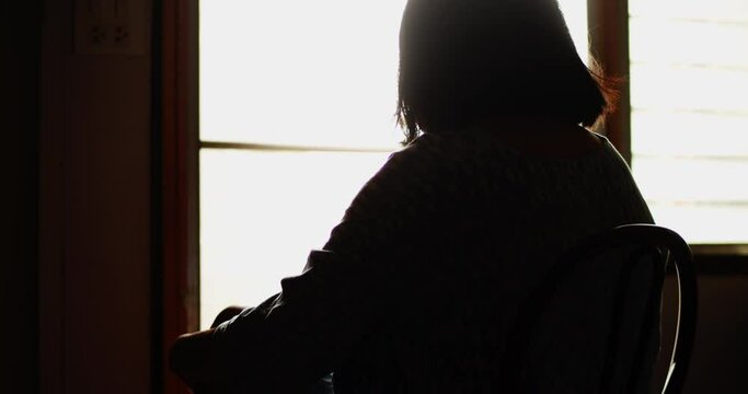 Silhouette Slow motion scene of a poor lonely depression woman sitting motionless thinking while swaying her body, looking out the door in the cramped room of her rented apartment.