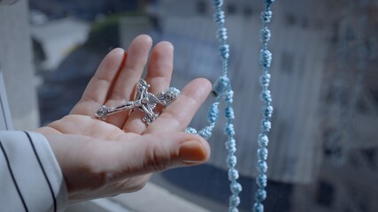 Tilt from close on hand holding crucifix to woman's hand counting through knots on a blue string rosary with blue sky in background