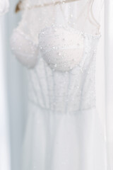 White wedding dress hanging near open window. The bride dress is hanging. Embroidery with pearls and beads.
