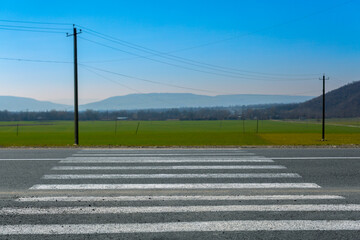 Crosswalk on the countryside road with a green field in the background, safe sidewalk with the pedestrian crossing. View on the field with mountains and crosswalk on the asphalt.