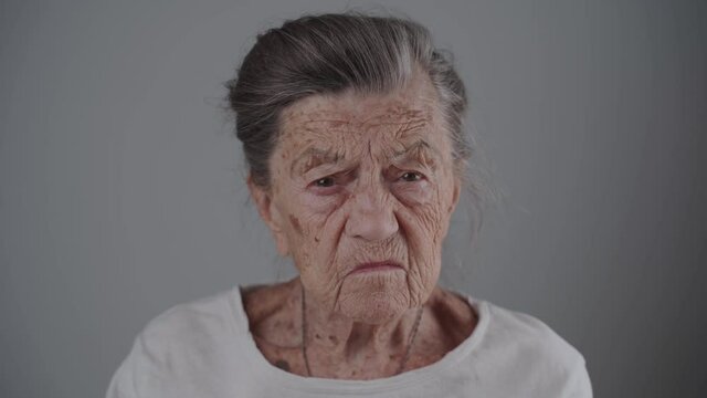 Portrait of an angry senior Caucasian woman with gray hair and deep wrinkles looking at camera on gray background. Very old female 90 years old negative emotion in the studio. Warning grandmother
