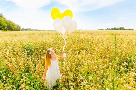 Happy child plays with white and yellow balloons outdoors. Kid having fun in a green spring field with flowers on a background of blue sky. Freedom and imagination concept.