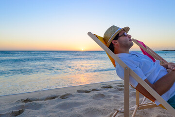 Young man relaxing on a beach chair with beach hat at sunset