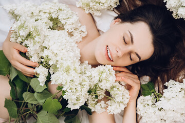 Obraz na płótnie Canvas Close up portrait of charming beautiful young woman with closed eyes and sincere smile lying in comfy white bed, spring flowers around head. Relaxed and cute girl with clear skin enjoys, feels joy