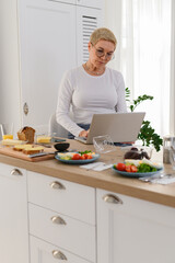 Senior woman working at laptop in kitchen. Looks cooking recipes online.